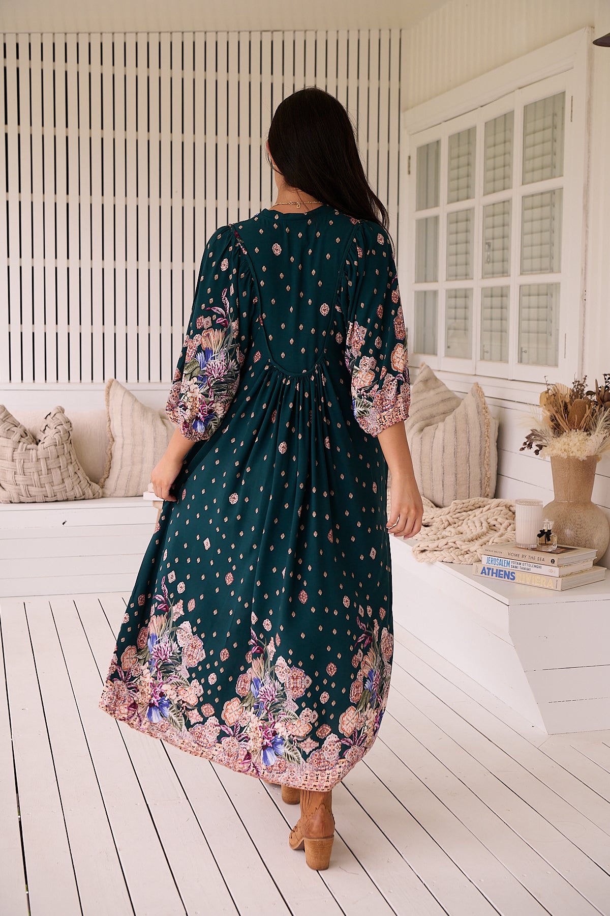 A woman standing in a room with white panelled walls, wearing a floral bohemian midi dress with a dark green base and long sleeves.
