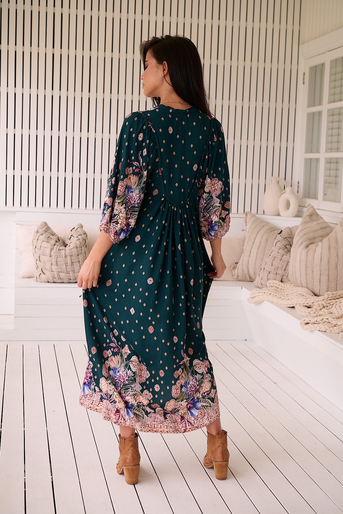 A woman standing in a room with white panelled walls, wearing a floral bohemian midi dress with a dark green base and long sleeves.