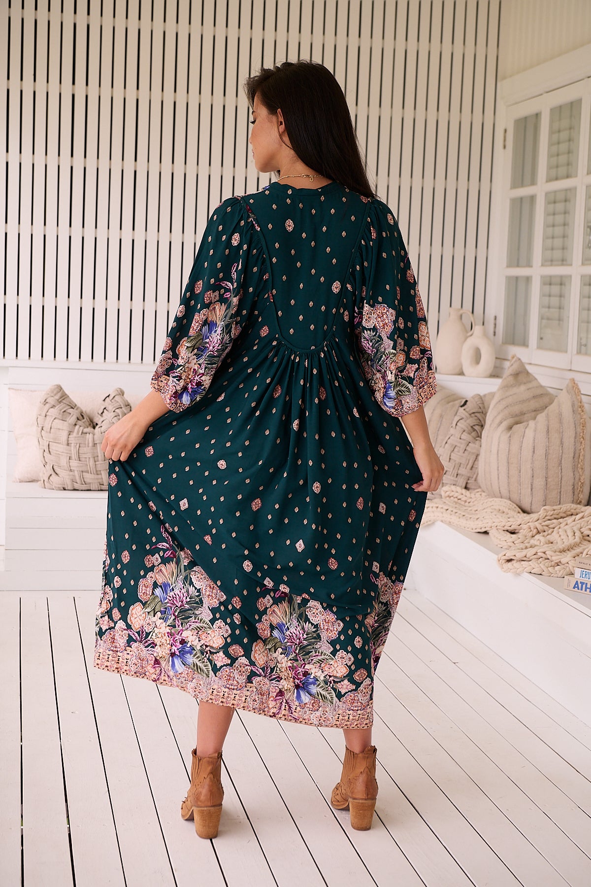 A woman standing in a room with white panelled walls, wearing a floral bohemian midi dress with a dark green base and long sleeves.