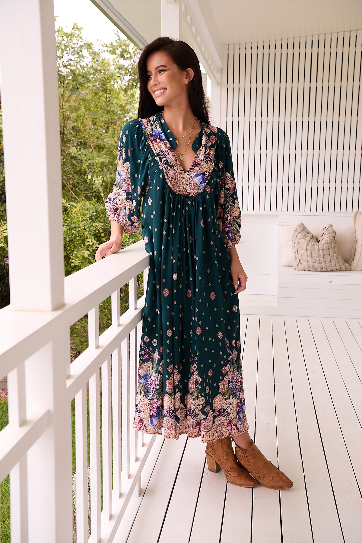 A woman standing in a room with white panelled walls, wearing a floral bohemian midi dress with a dark green base and long sleeves.