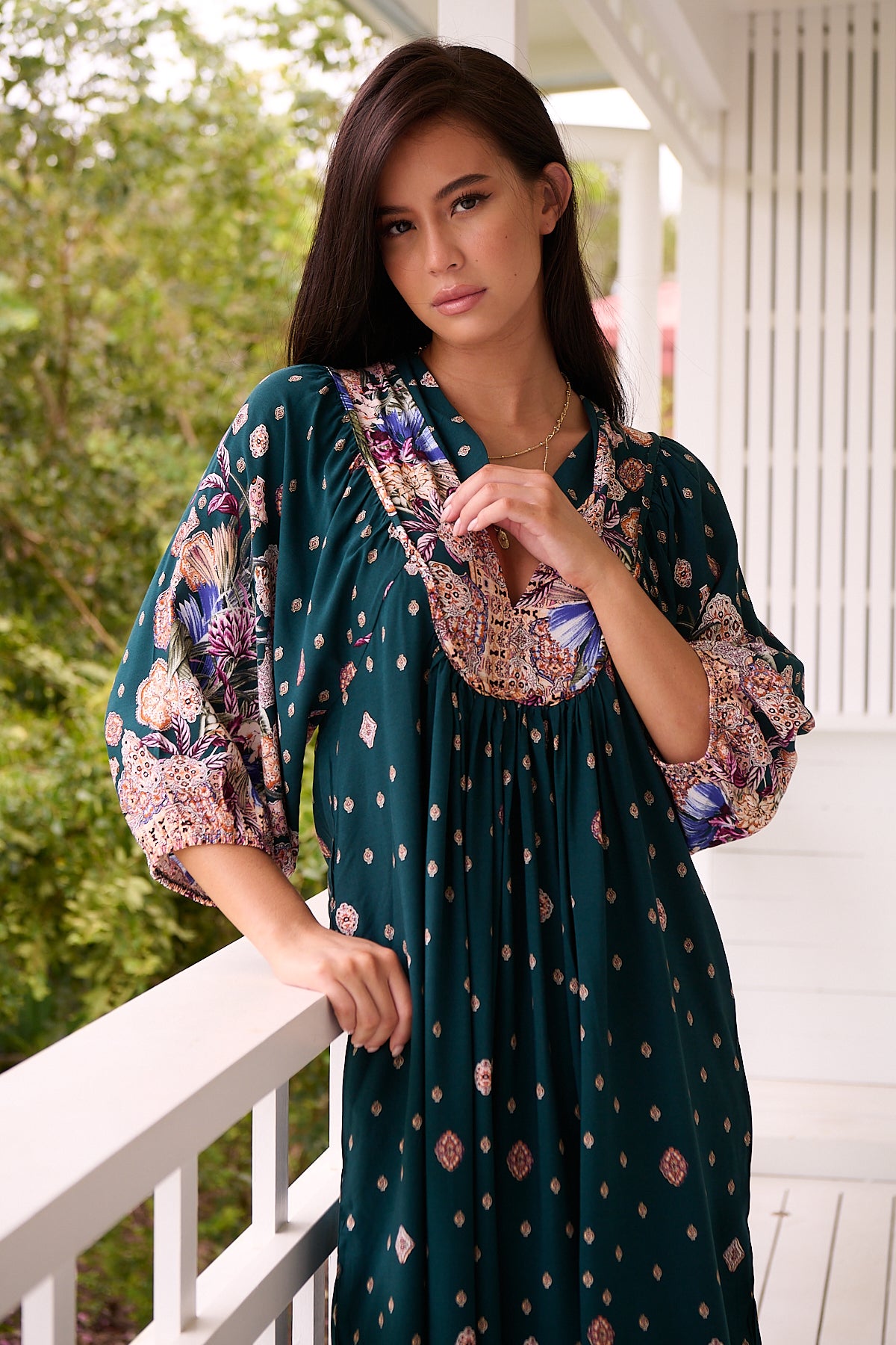 A woman standing in a room with white panelled walls, wearing a floral bohemian midi dress with a dark green base and long sleeves.