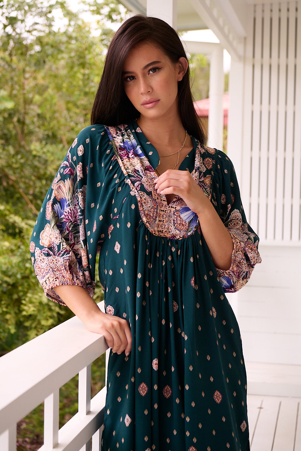 A woman standing in a room with white panelled walls, wearing a floral bohemian midi dress with a dark green base and long sleeves.