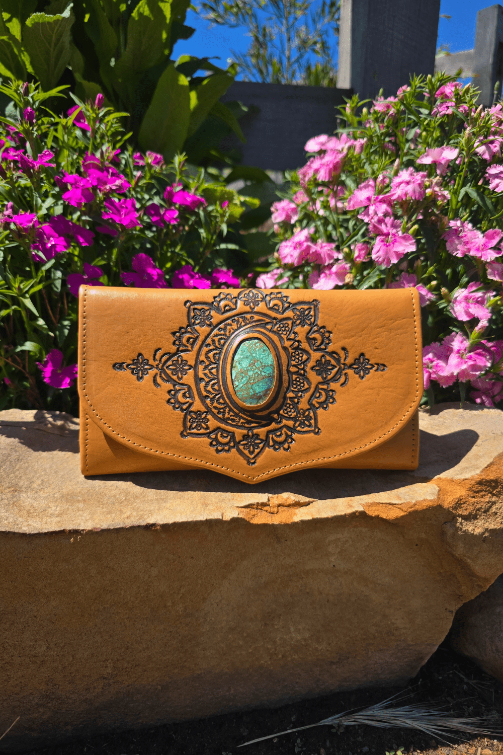 A light tan leather wallet with a hand-tooled crescent moon, mandala, and floral patterns, placed on a stone surface with flowers in the background.