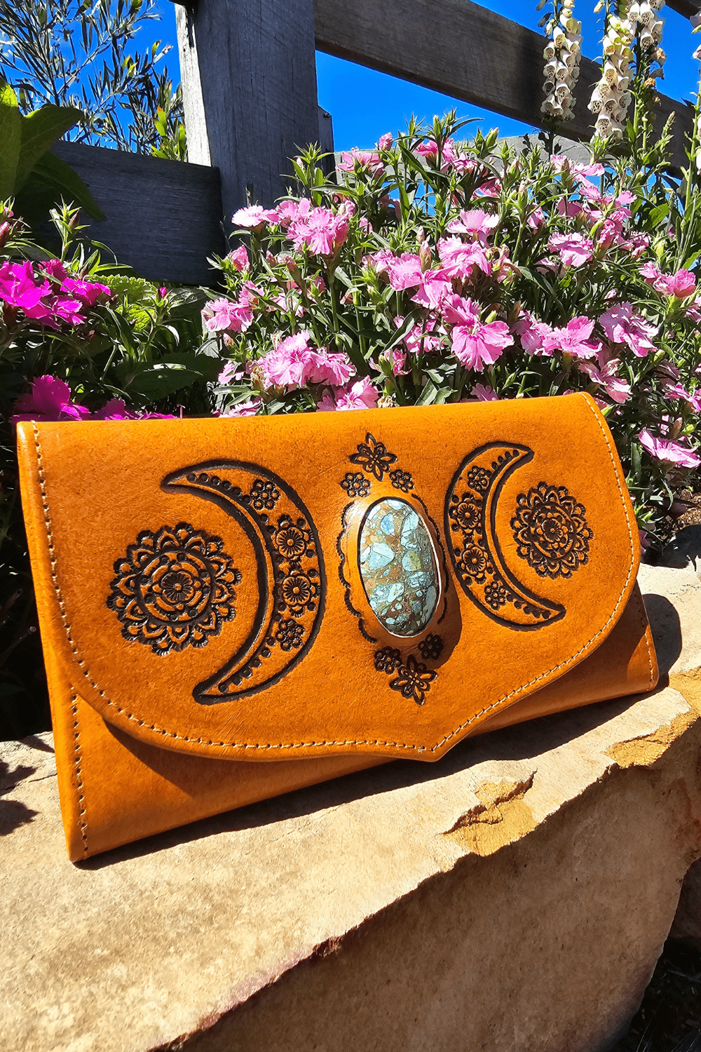 A light tan bohemian leather wallet with hand-tooled crescent moons and floral patterns, displayed on a stone surface with flowers in the background.