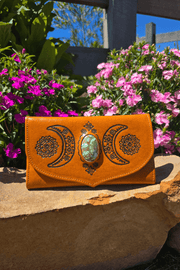 A light tan bohemian leather wallet with hand-tooled crescent moons and floral patterns, displayed on a stone surface with flowers in the background.