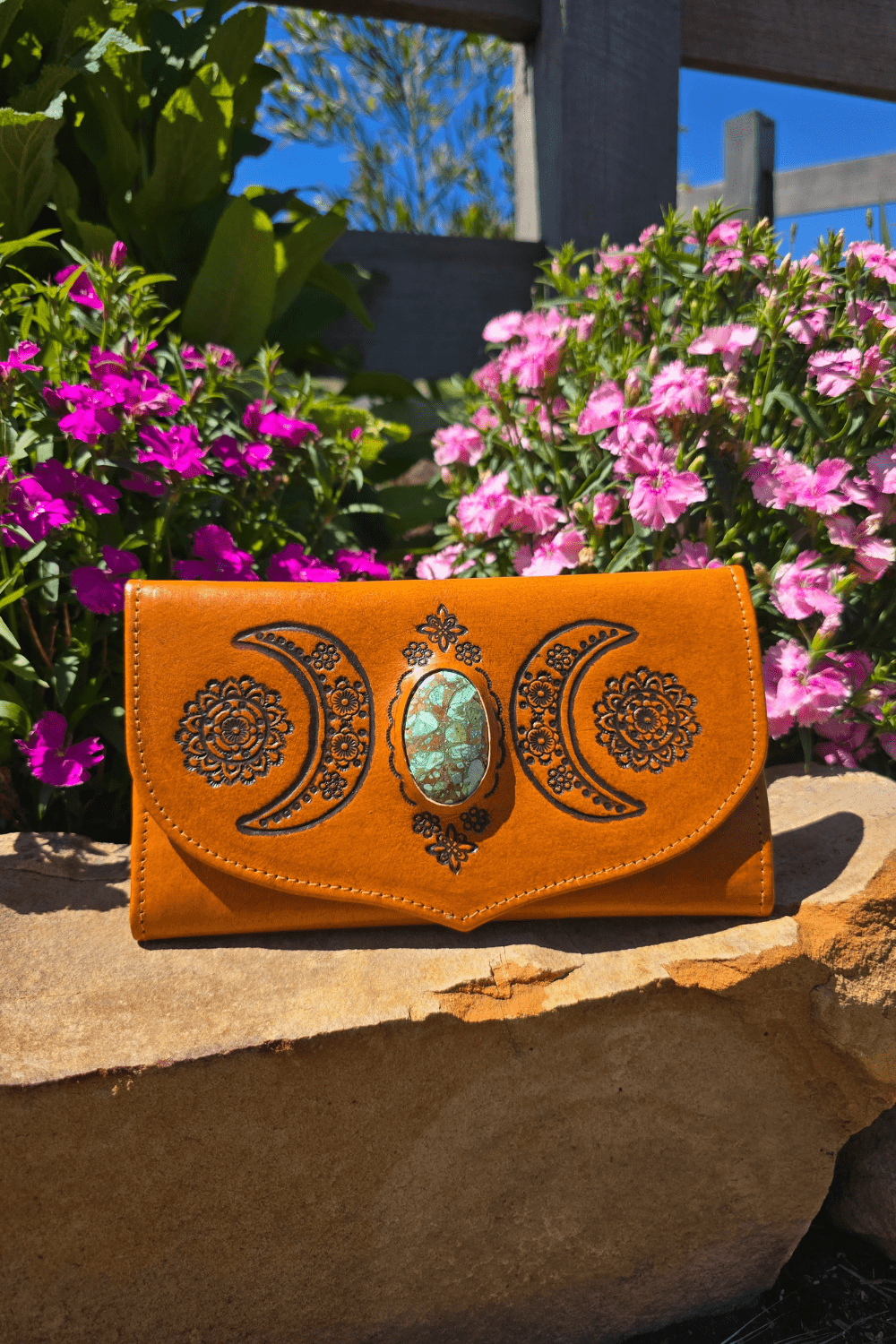 A light tan bohemian leather wallet with hand-tooled crescent moons and floral patterns, displayed on a stone surface with flowers in the background.