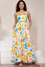 A woman wearing a white maxi dress with a vibrant multicolored floral print. The dress features a straight square neckline, thick shoulder straps, a shirred back panel, and a tiered skirt.
