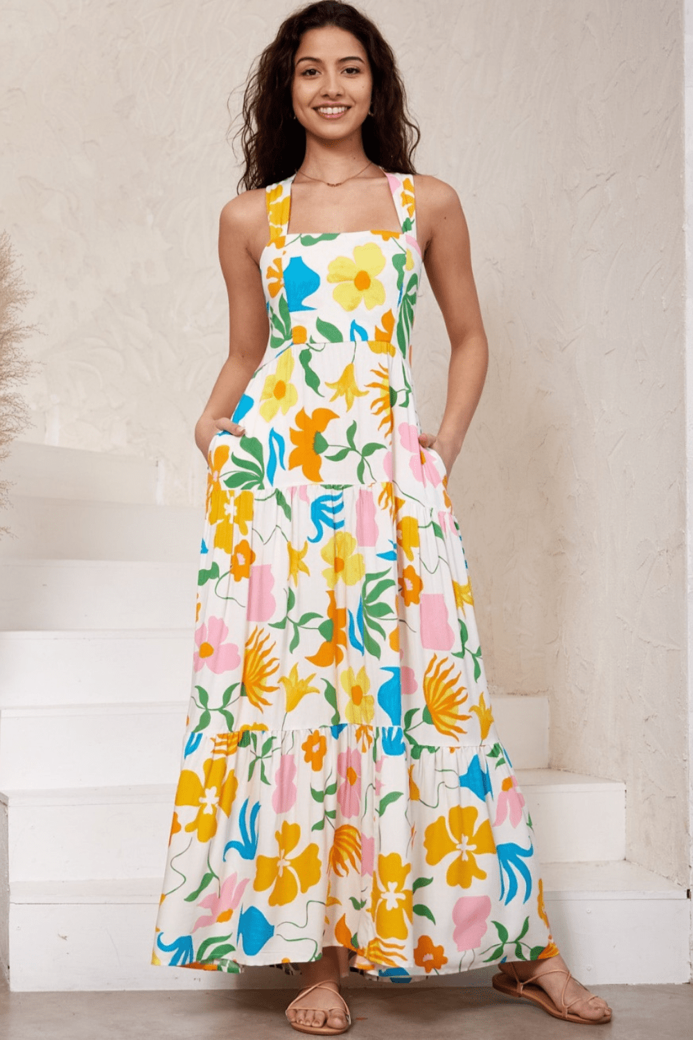 A woman wearing a white maxi dress with a vibrant multicolored floral print. The dress features a straight square neckline, thick shoulder straps, a shirred back panel, and a tiered skirt.