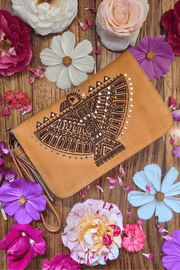 A tan leather wallet with an intricately laser-etched spirited eagle design, displayed on a surface with scattered flowers and a zippered coin pouch visible at the bottom.