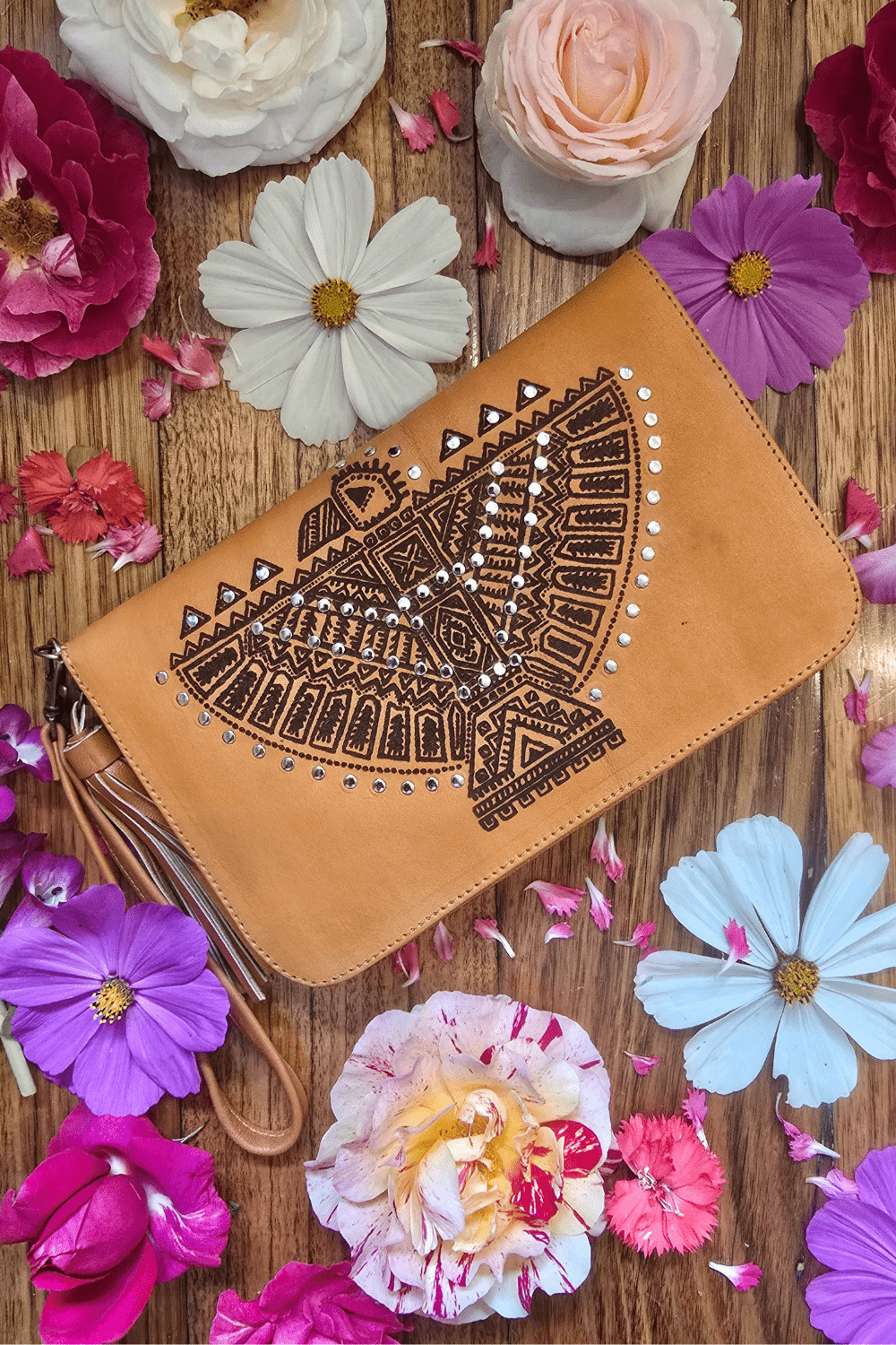 A tan leather wallet with an intricately laser-etched spirited eagle design, displayed on a surface with scattered flowers and a zippered coin pouch visible at the bottom.