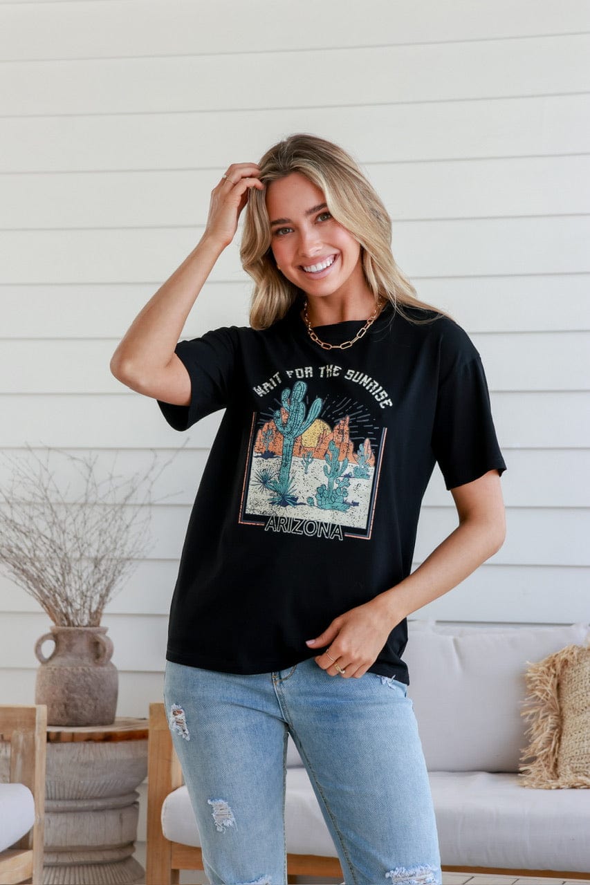 Woman smiling modelling a black cotton graphic tee with Cacti in the desert and the slogan 'Wait for the sunshine, Arizona'